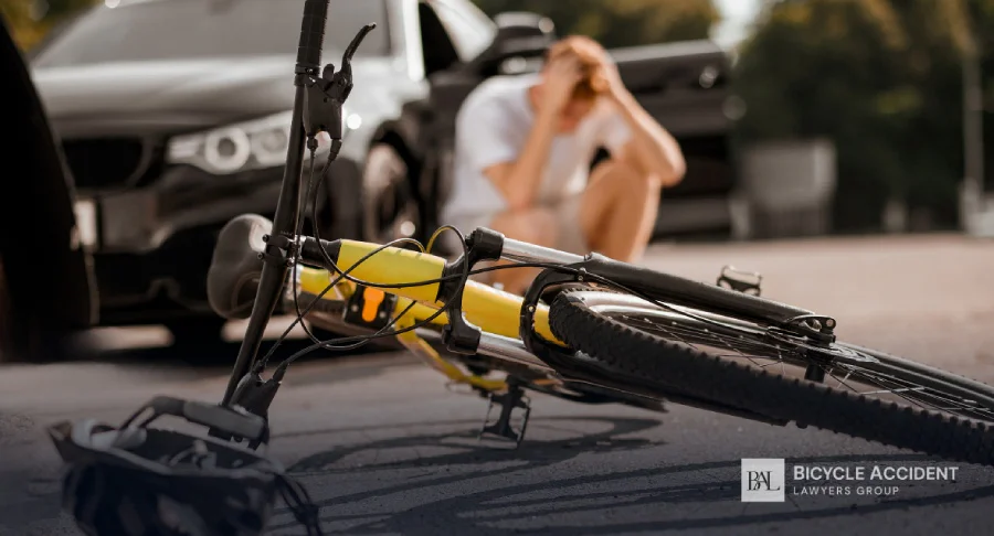 A yellow bicycle lies on the road near a car with a distressed man sitting nearby.