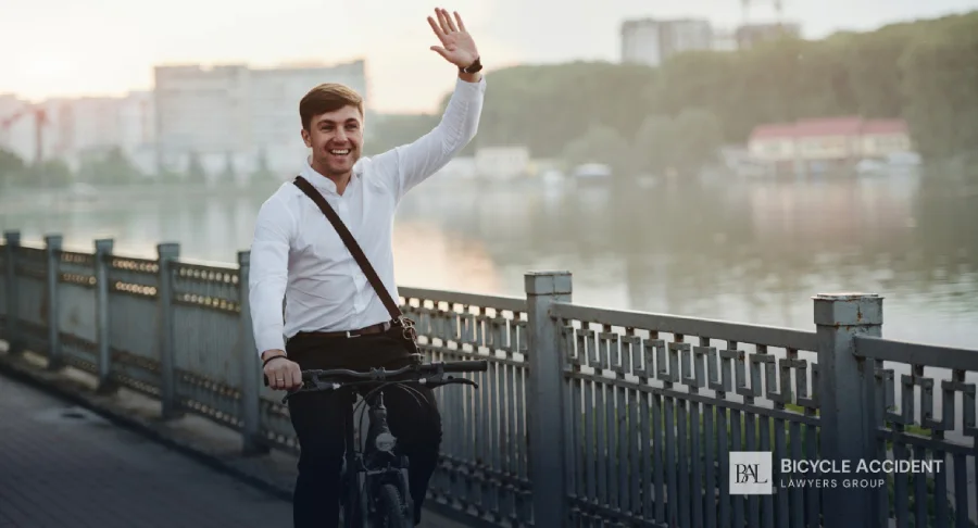 A smiling man in a white shirt waves while riding his bicycle along a scenic waterfront.