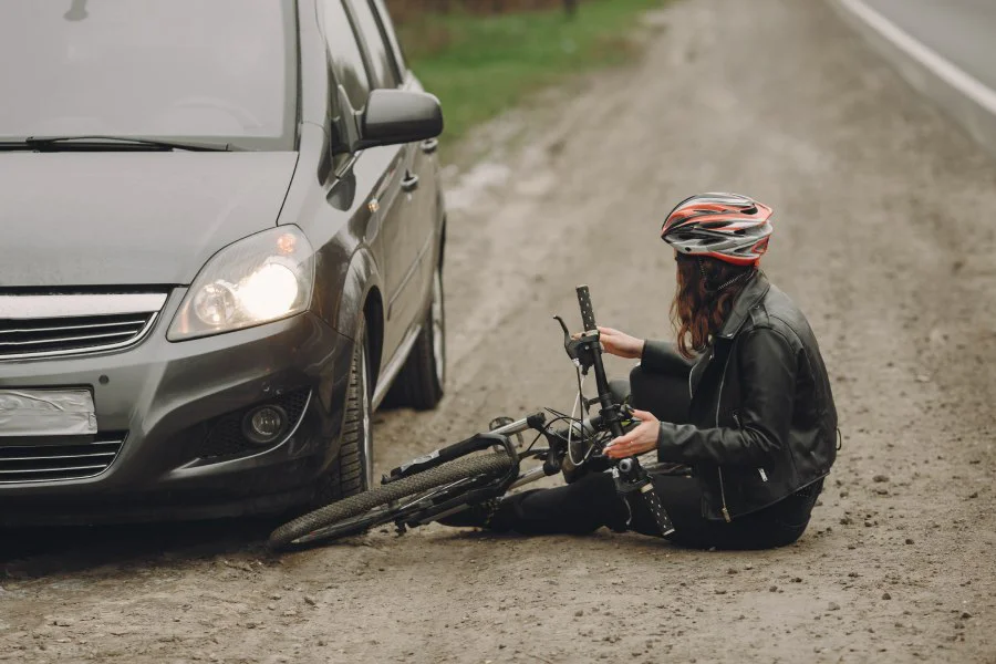 A cyclist in a helmet sits by her bike after a collision with a grey car.