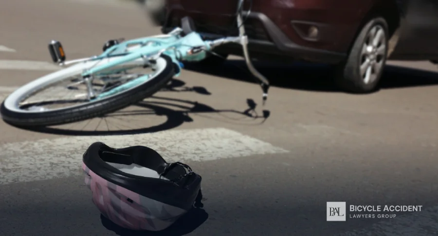 A pink bicycle helmet sits on the asphalt in front of a fallen light-blue bike and car.