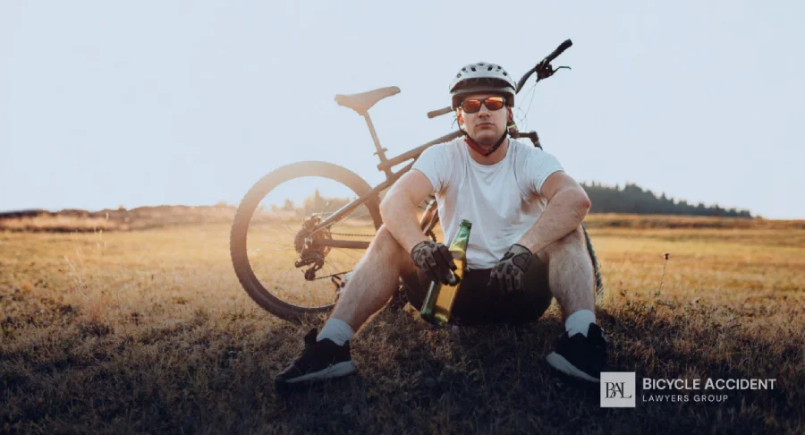 A cyclist in a white shirt and helmet sits on the grass holding a glass bottle.