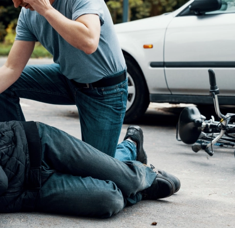 Man helping an injured cyclist on the road next to a fallen bicycle and car.